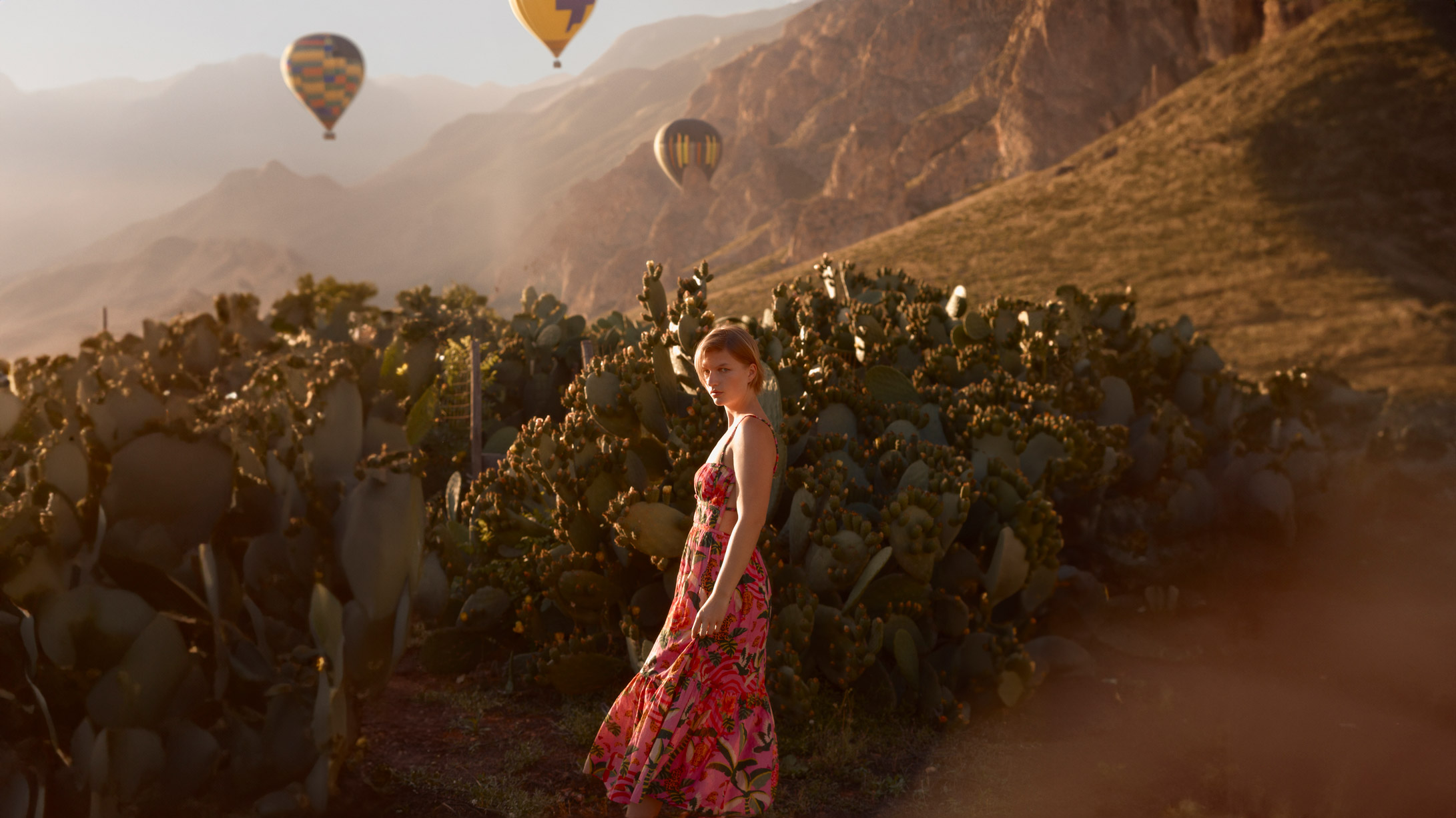 Photographie d'une femme en robe rose devant un paysage de montgolfiere, de montagnes et catcus